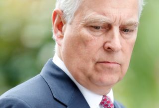 Prince Andrew wearing a blue shirt, white shirt and red tie looking down