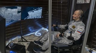 a man in a gray jumpsuit sits in a room with computer screens showing lunar landscapes