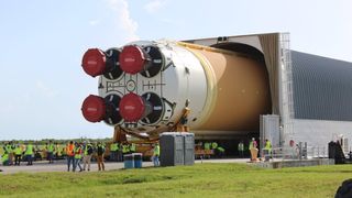 a big rocket on its side emerges from a storage facility. four engines are visible with plastic covers on the bells. a lot of people in green and orange construction photos stand nearby