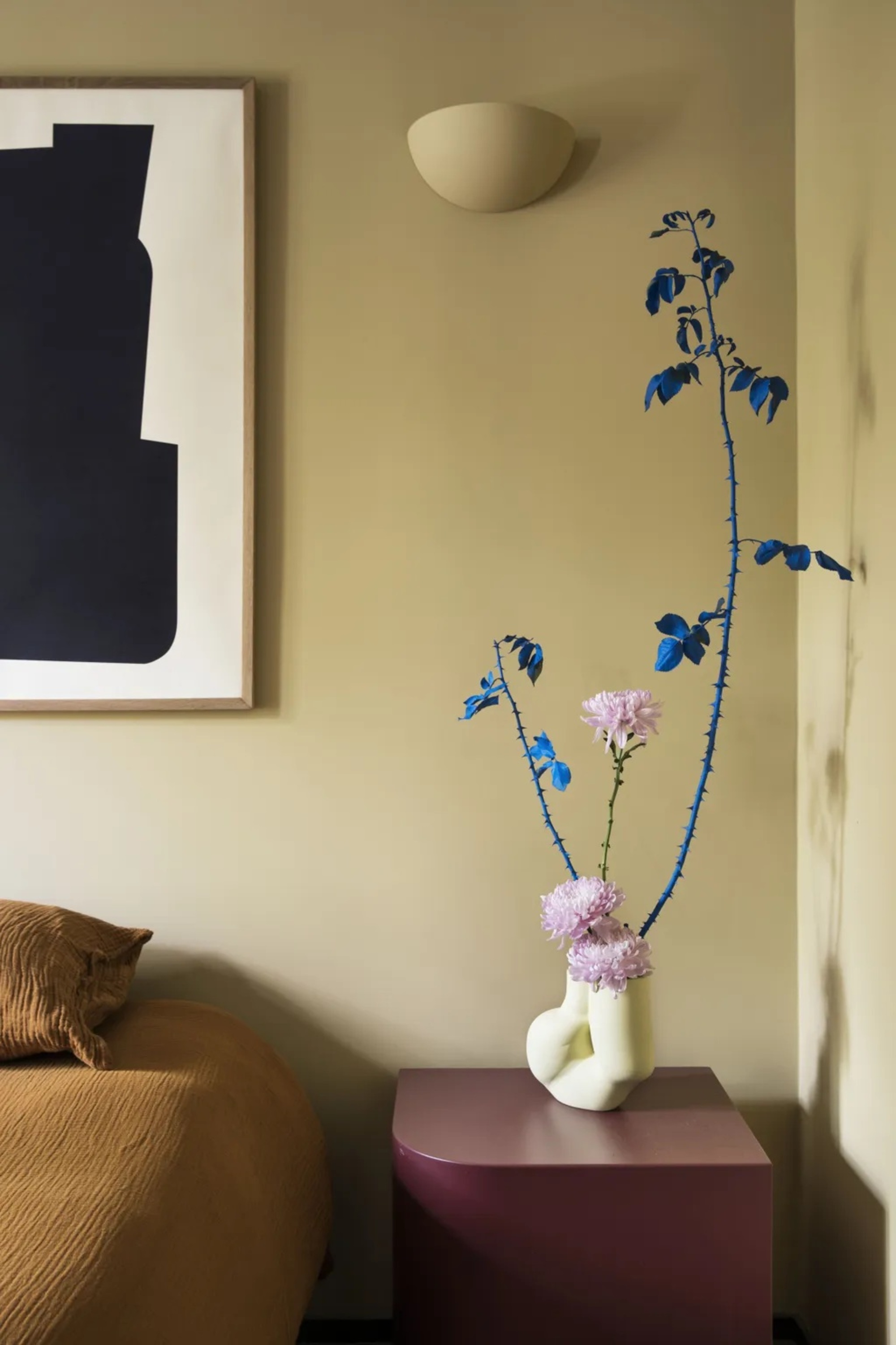 close up of bedroom with butter yellow walls, mustard yellow bed linen with artwork hanging above bed, burgundy side table with small yellow vase with stem painted blue with pink flowers