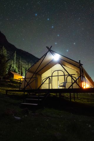 a large tent is illuminated from within, there are steps leading to the entrance. Above is a starry sky with a distinct pattern of stars known as the big dipper shining bright.