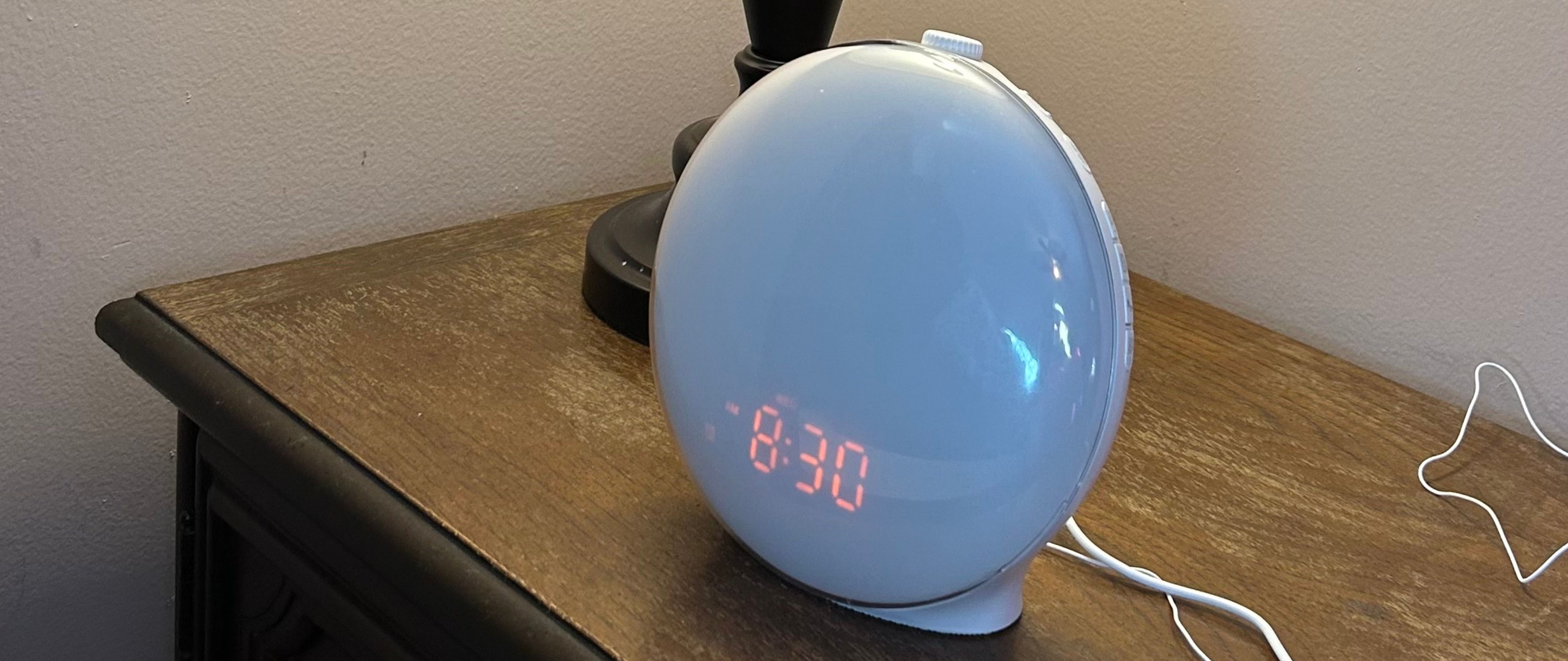 The white JALL Wake-Up Light Sunrise Alarm Clock with digital time display on wooden bedside table.