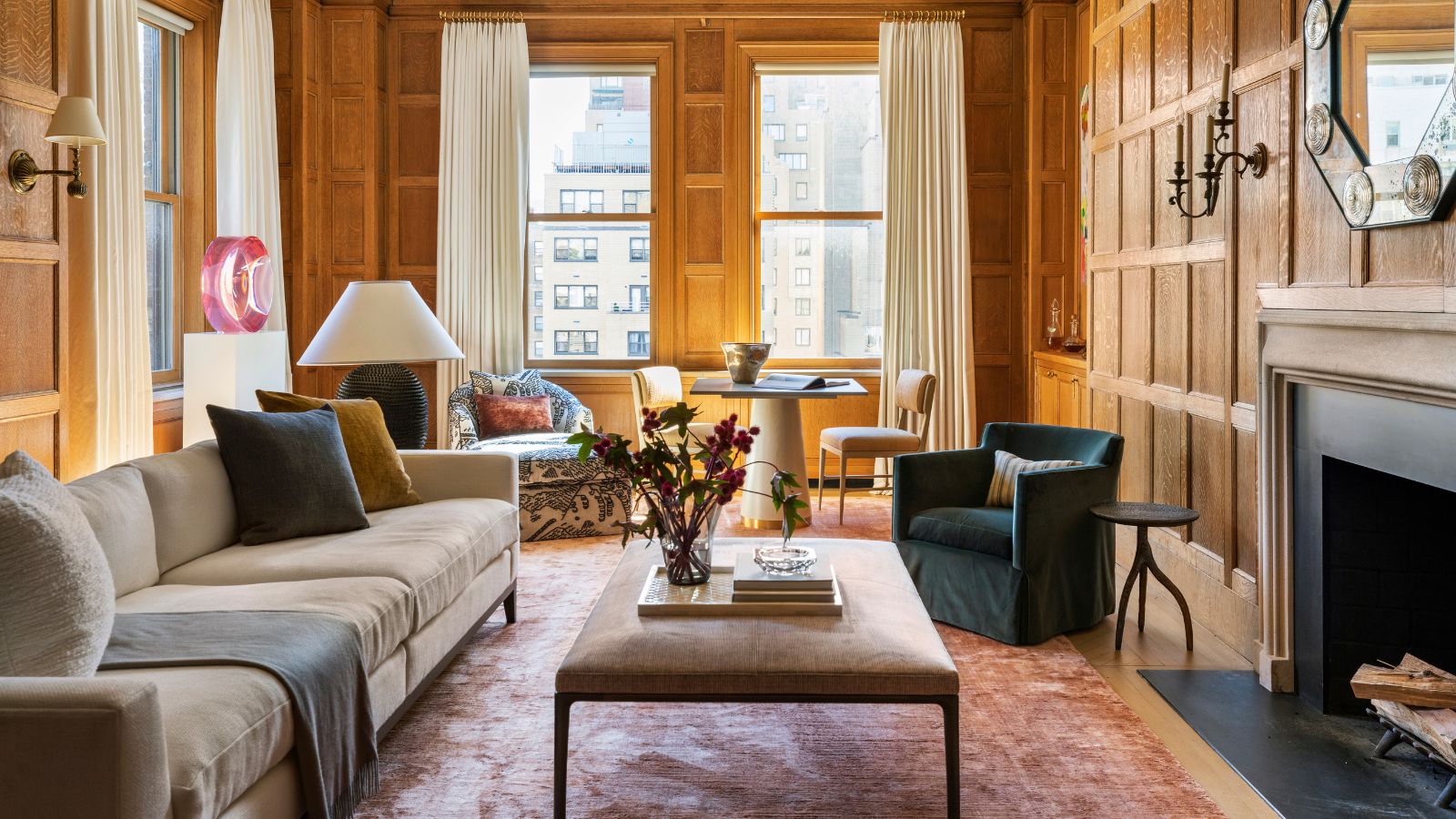 large cozy living room with original wooden panelled walls and fireplace with a large rug, beige sofa, ottoman, and a little seating area with table and chairs