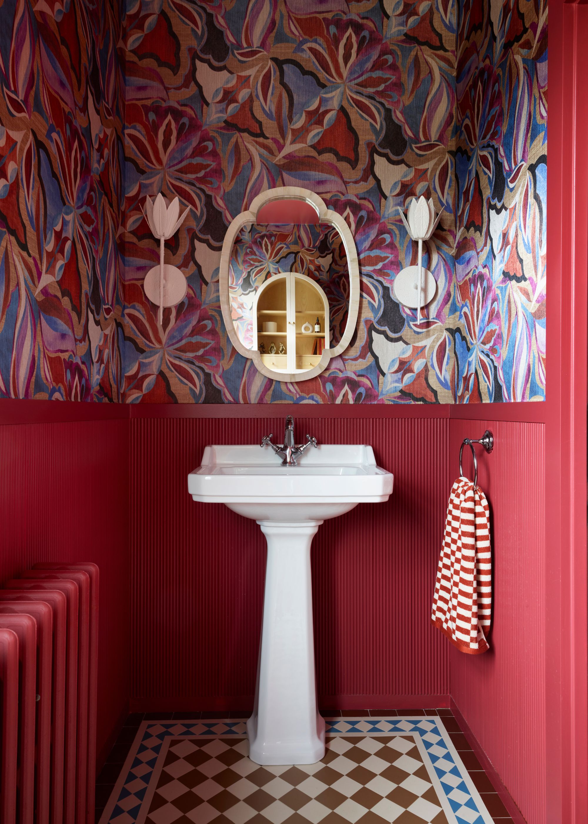 A powder room with red reeded wall paneling on the lower half of the walls and colorful, patterned wallpaper above. Geometric floor tiles and a white sink and decorative wall mirrors, and sconces.