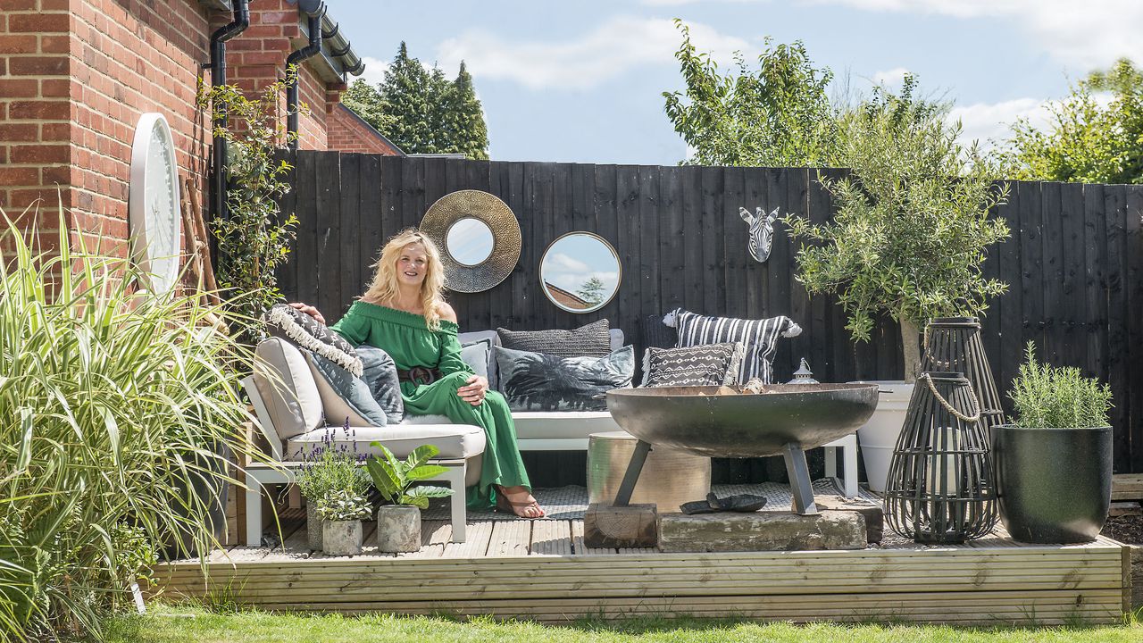 Woman sitting on outdoor corner sofa with firepit and dark fence