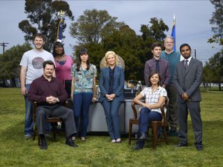 the cast of parks and recreation poses together around a desk on a field