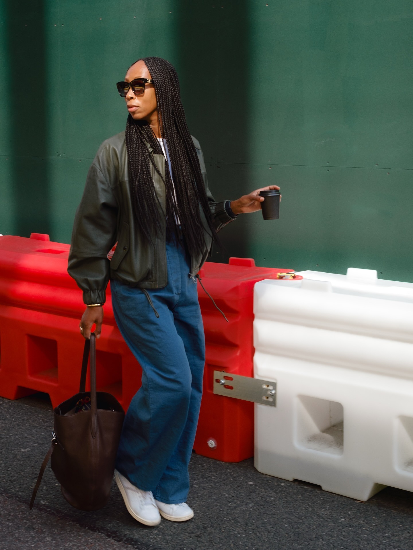 Woman wears green leather jacket, blue trousers, white trainers and carries brown bag