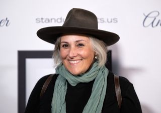 ricki lake smiles and poses on a red carpet in a black wide brimmed hat and green scarf promoting The High Life