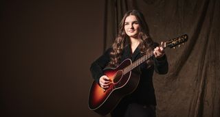 Muireann Bradley wears black and plays her custom-built sunburst acoustic as she is photographed against a dark brown background.