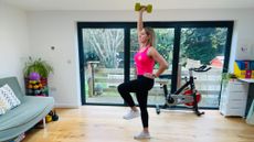 Personal trainer Maddy Biddulph performs a weighted dumbbell march. She wears a vest, leggings and sneakers and exercises in a living room with a wooden floor and white walls. She is standing on her right leg with her left leg off the floor and bent at the knee. Her left hand is on her hip while her right hand grips a dumbbell overhead. Behind her we see an exercise bike and sliding glass doors leading to a garden.