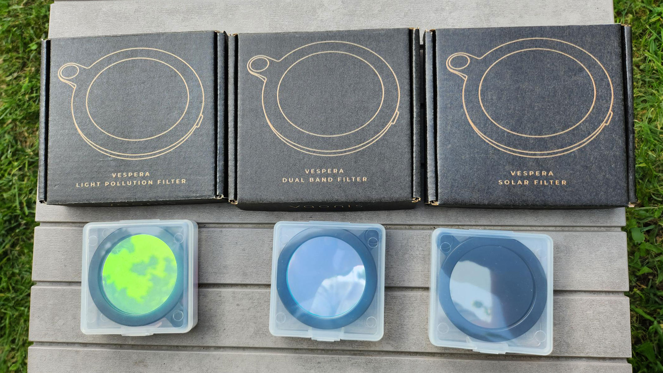 The Vespera Light Pollution Filter, Vespera Dual Band Filter and Vespera Solar Filter with boxes on a grey table with grass at the side.