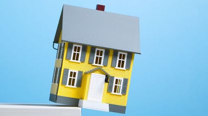 Miniature model of yellow single family house about to fall off edge of white shelf, blue background
