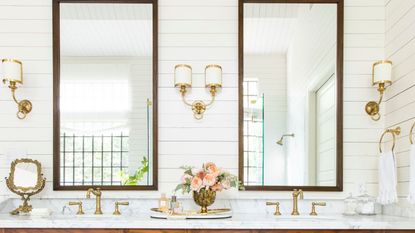 Bathroom with Cloud White painted panelled walls, large mirrors with wooden frames, a marble vanity unit with double sinks and gold taps, and gold and white sconces flanking the mirrors