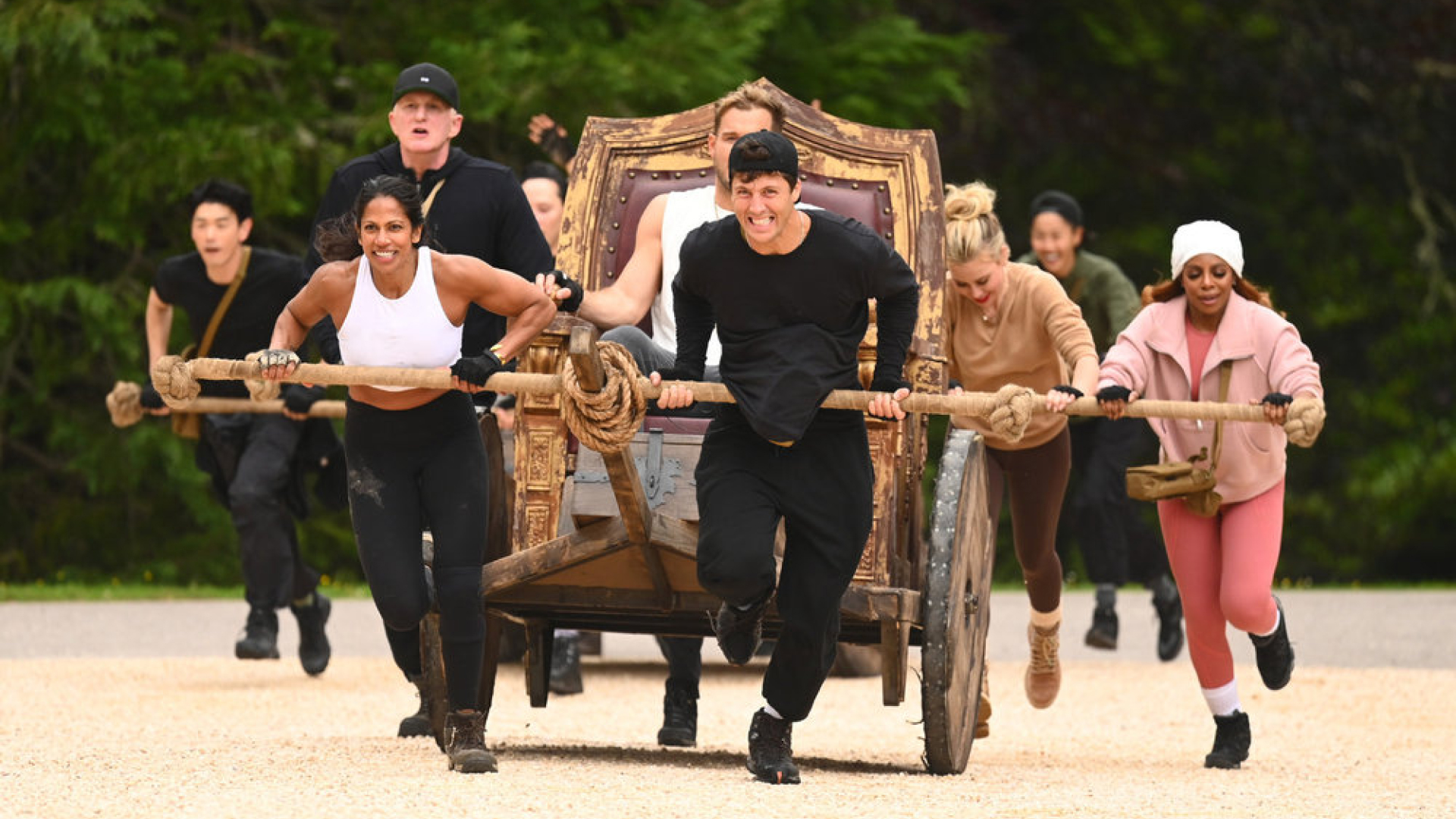 A group of people (l-r Eric Nam, Natalie Anderson, Michael Rapaport, Rob Rausch, Tara Lipinski, Candiace Dillard Bassett) push a throne-chariot with a man (Colton Underwood) sitting on it, in 'The Traitors' season 4.