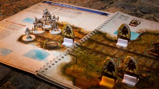 Multiple player hero miniatures stand on the Gloomhaven: Jaws of the Lion scenario book along with cardboard monster standees