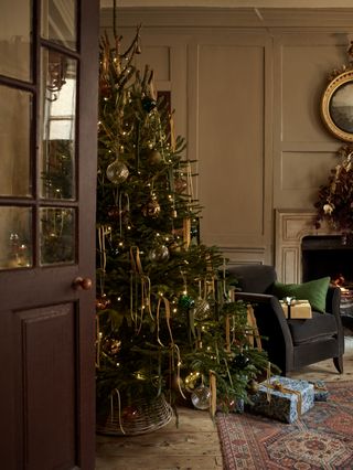 Christmas living room with stone colored walls, real tree with gold ribbon, grey armchair with presents, open fire, antique rug