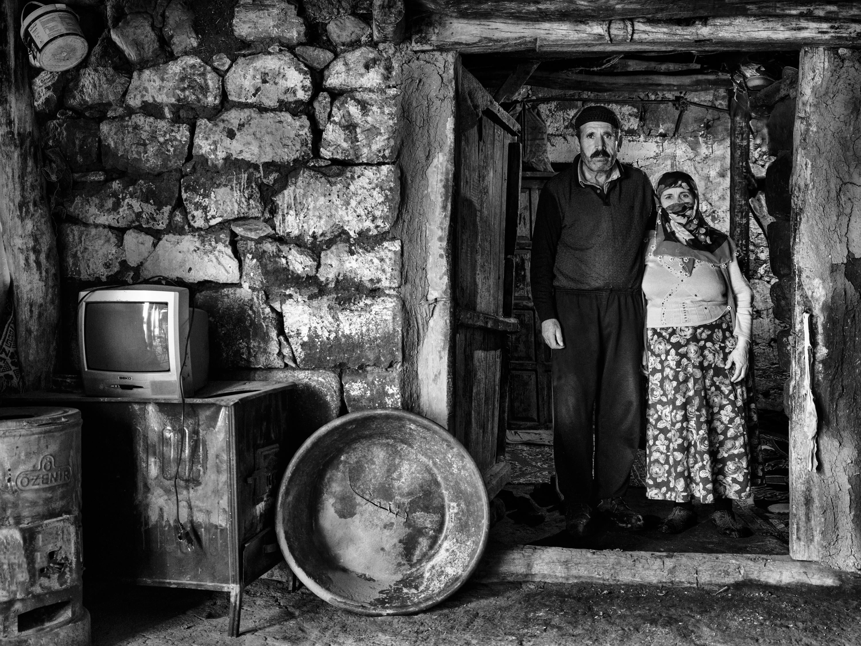 Black and white portrait of a man and woman standing together in a rustic stone doorway, beside an old television set and a large dented metal basin.