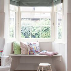 Bay window seat in a large bay window in a white room, covered in cushions and throws