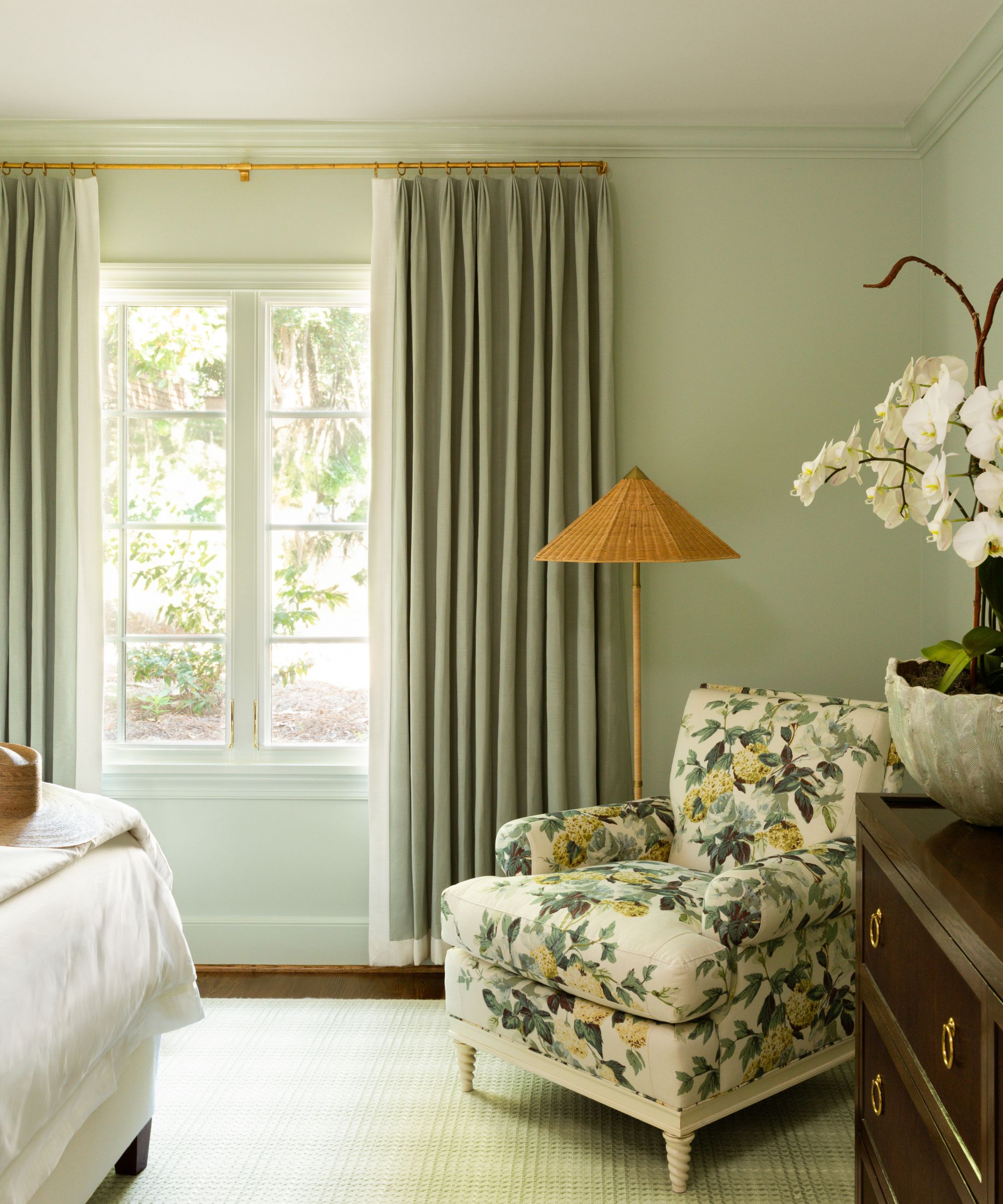 A calming bedroom painted light green with green curtains, a green and yellow floral armchair, a woven floor lamp, a dark wooden dresser, and a pale green rug.