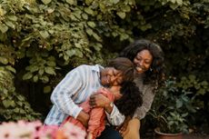 A grandparent hugs her grandchild with her mother smiling outside by trees.