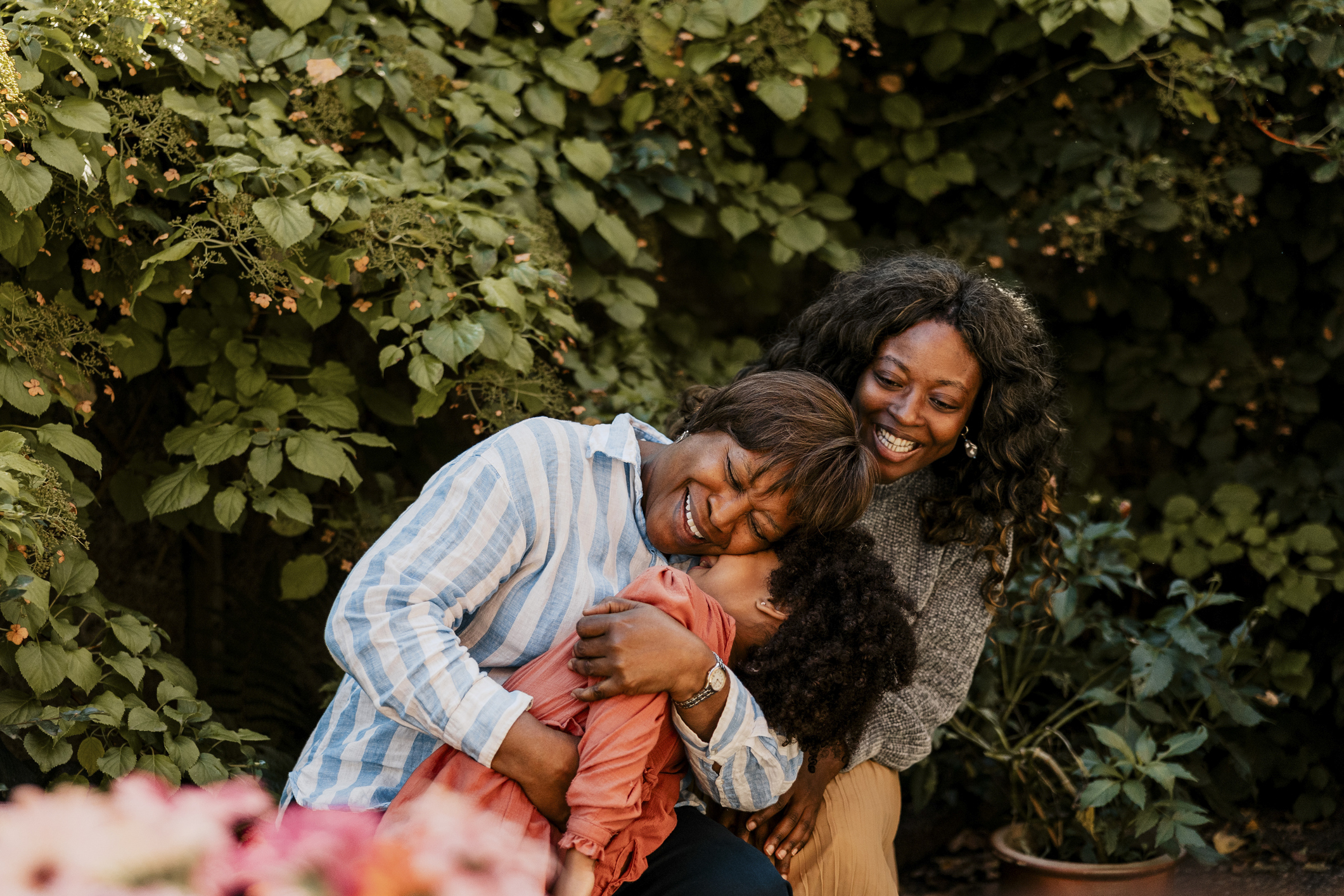 A grandparent hugs her grandchild with her mother smiling outside by trees.