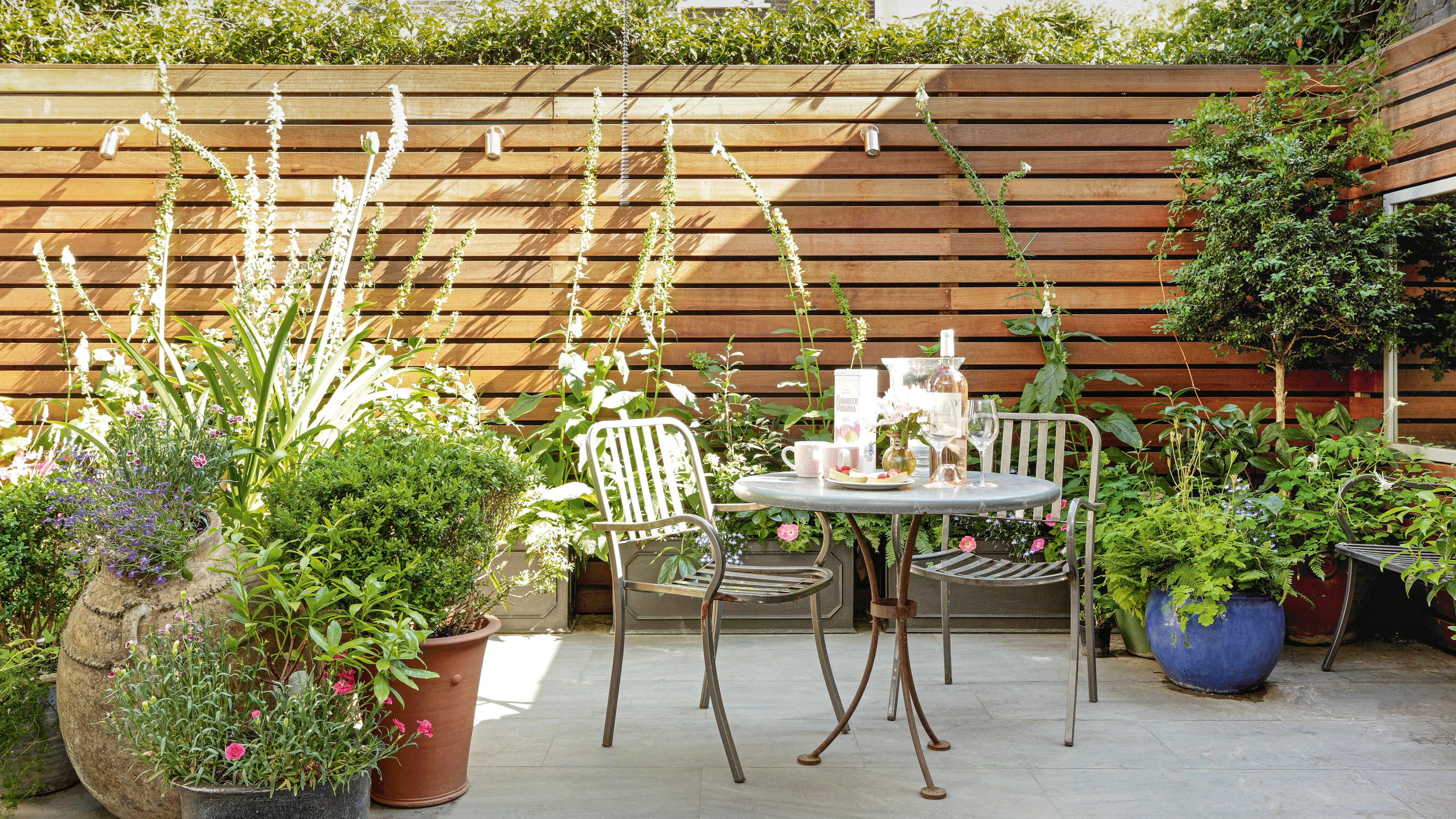 Table and chairs laid for a meal on a gravel area beside raised beds with shrubs and flowers. 