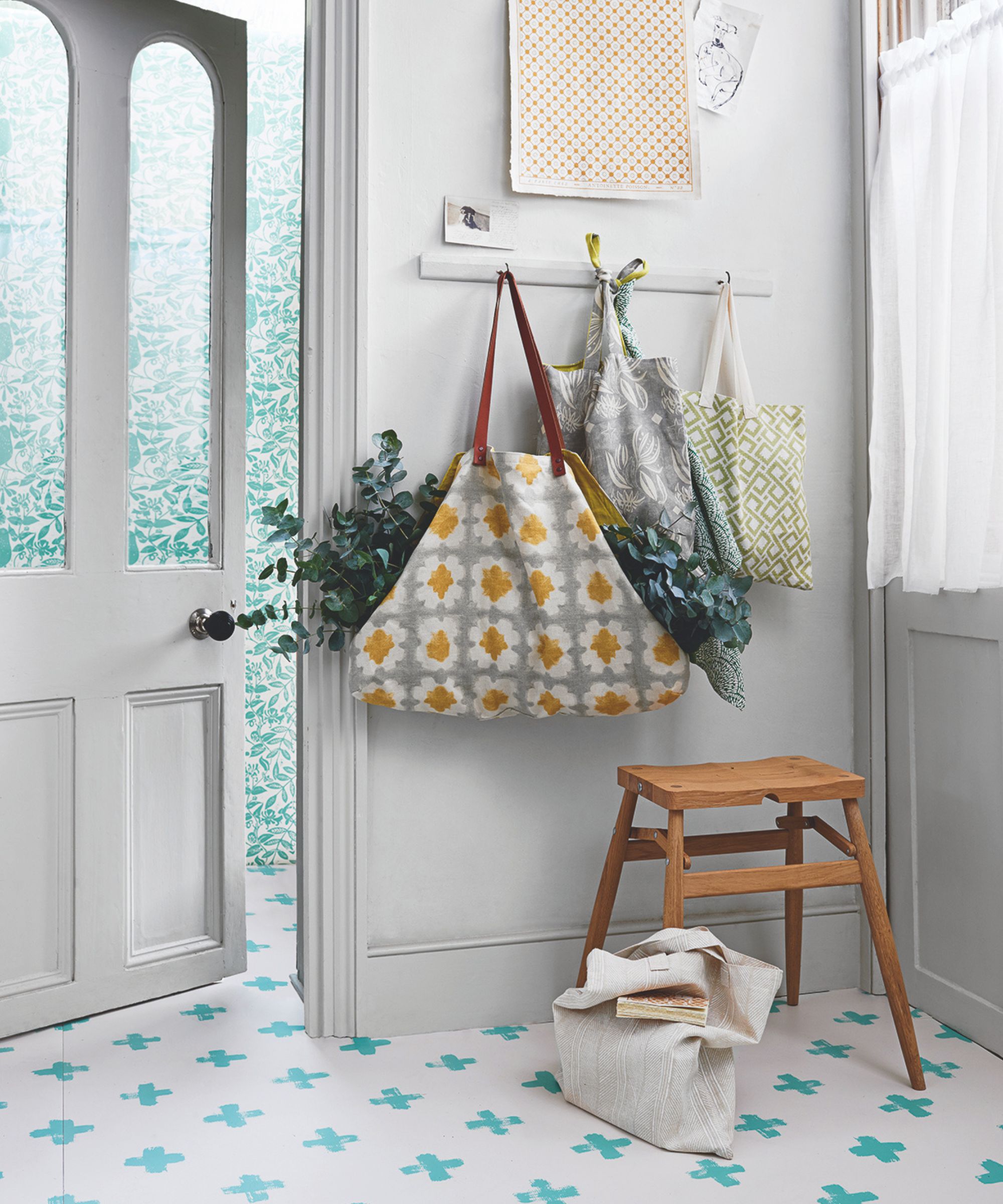 A white entryway space with blue and white tiled flooring, a white glass-paned door and wall hooks with bags filled with plants. Underneath them is a wooden stool, and to the right is a window with a sheer curtain.