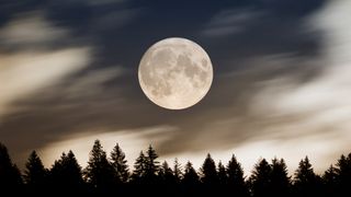 A full moon rises over silhouetted conifers, against a dark sky smeared with blurry wisps of clouds.