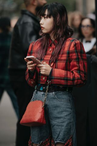 A woman at fashion week wearing a flannel shirt outfit featuring a red cropped jacket, blue jeans, and a red bag.