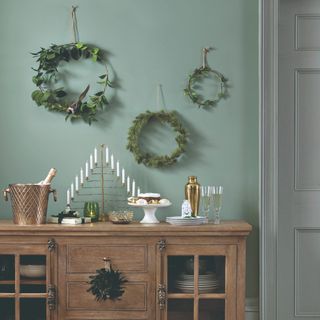 A dining room with a sideboard painted in mint green decorated with three simple Christmas wreaths hanging on the wall