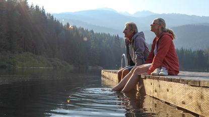 Mature couple relax on wooden pier, looks out across lake, BC, Canada.
