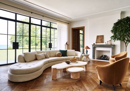A neutral living room with a curved white sofa, large French windows, wooden herringbone flooring and velvet orange chairs