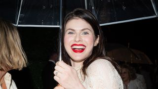 Alexandra Daddario shows off her wedding ring while holding an umbrella and smiling with red lipstick at the Dior show.