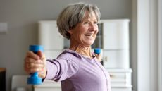 A smiling woman holds light dumbbells out to the sides with straight arms. 