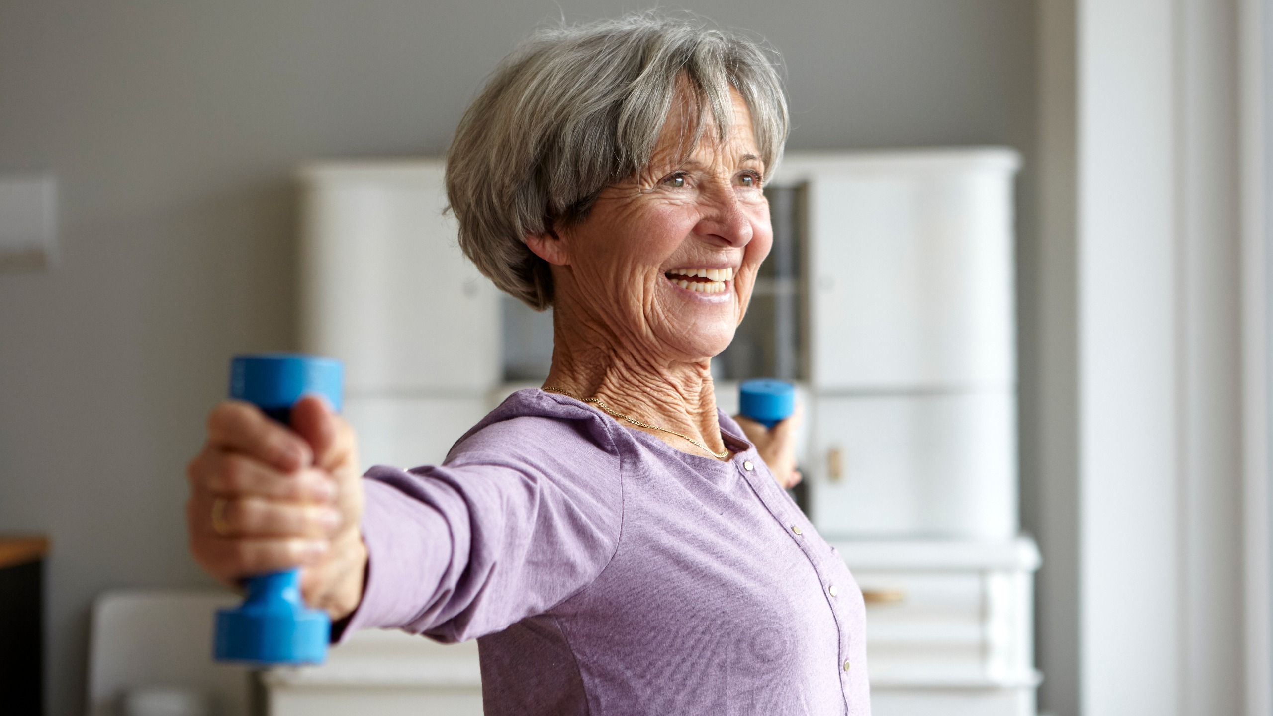 A smiling woman holds light dumbbells out to the sides with straight arms. 