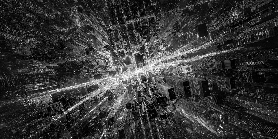 Black-and-white aerial view of a dense city at night, with illuminated streets radiating from a bright central intersection