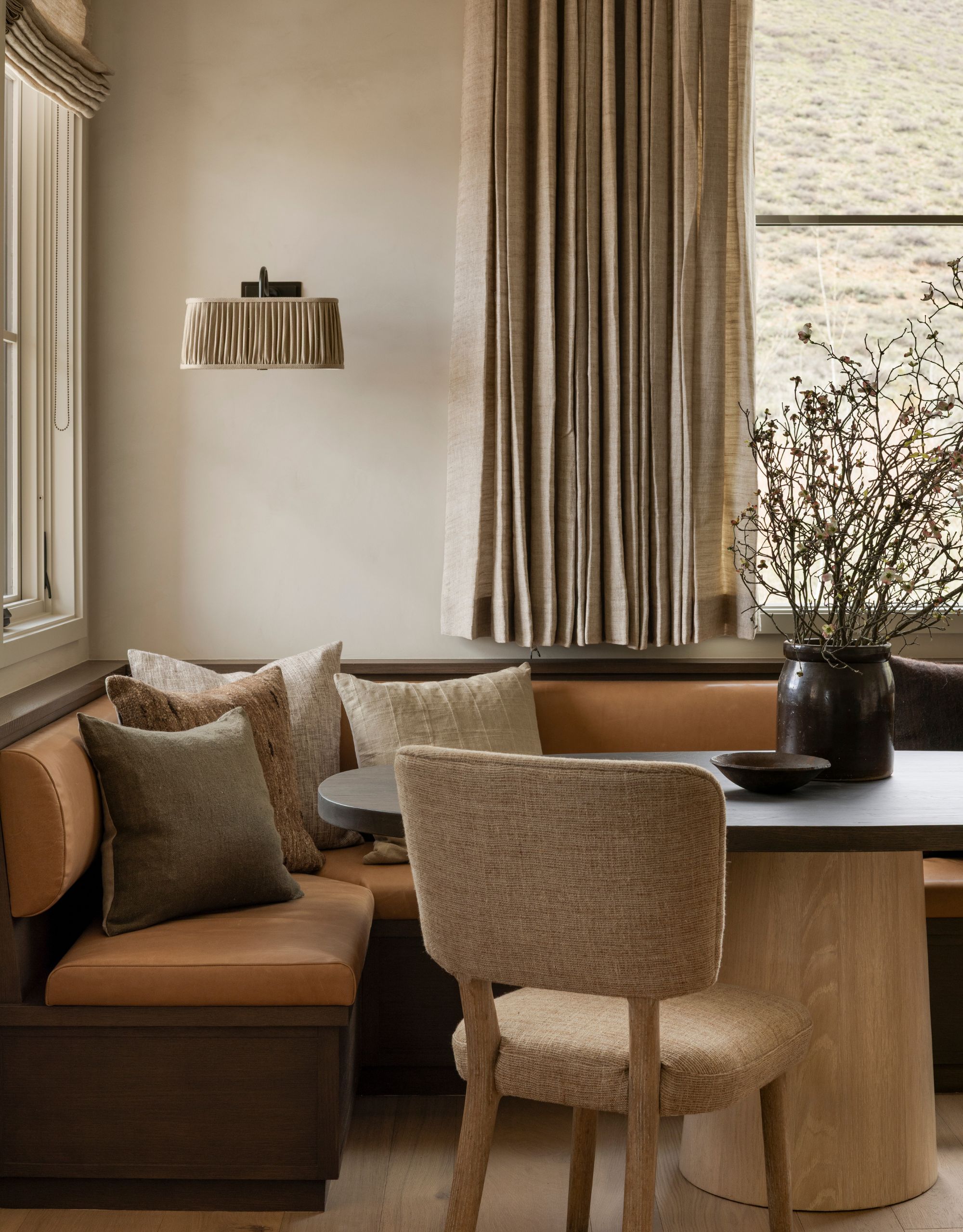 A modern corner banquette with orange seating, a black oval table top with a wooden pedestal base with cream walls and curtains and a modern pleated wall light sits above the corner of the banquette