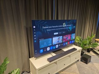 Samsung QN990F on a white media unit with a grey curtain in the behind it and soundbar in front