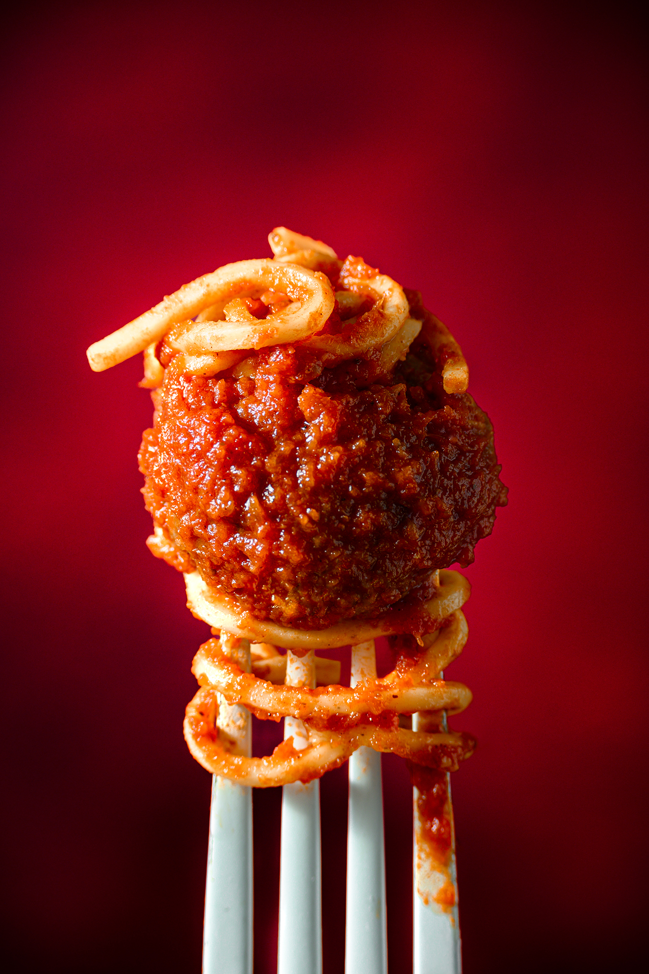 A fork holding a spaghetti-wrapped meatball drenched in red tomato sauce, against a vibrant red background