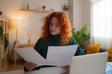 Young adult woman reviewing household bills and invoices, organizing personal finance while sitting at a desk with a laptop