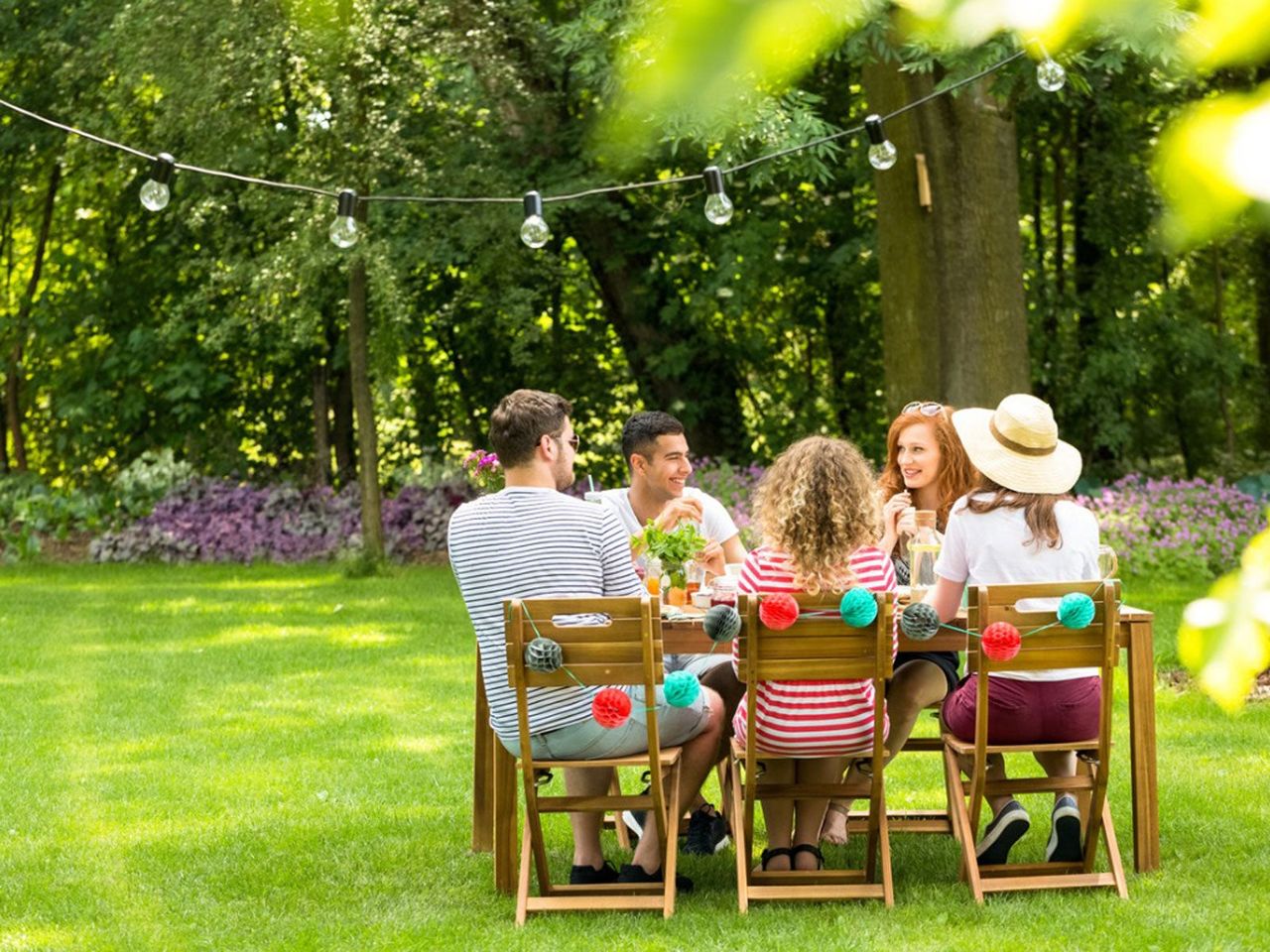 People Sitting At A Table On The Green Lawn With Trees And Flowers In The Background