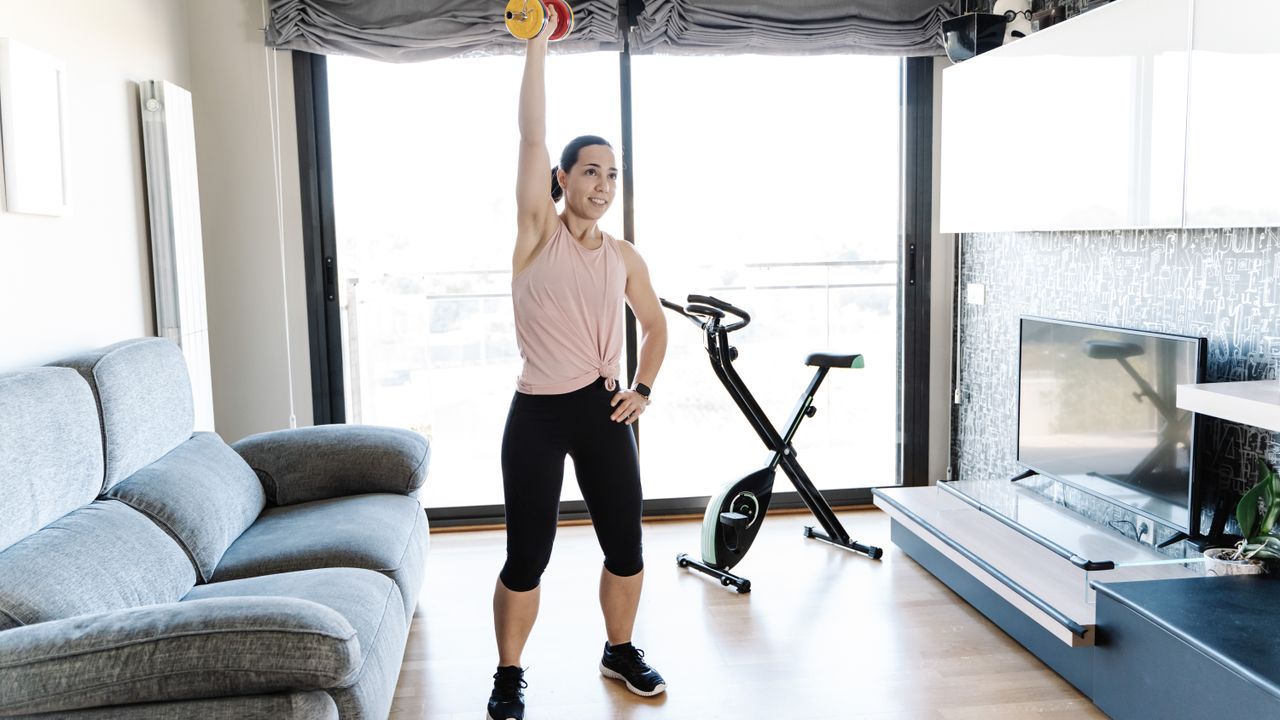 A woman exercises with a dumbbell at home. She is standing and holding the weight overhead, with a straightened arm; her other hand rests on her hip. Behind her is an exercise bike and large window. To either side of her is a couch and mounted television.