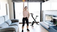 A woman exercises with a dumbbell at home. She is standing and holding the weight overhead, with a straightened arm; her other hand rests on her hip. Behind her is an exercise bike and large window. To either side of her is a couch and mounted television.