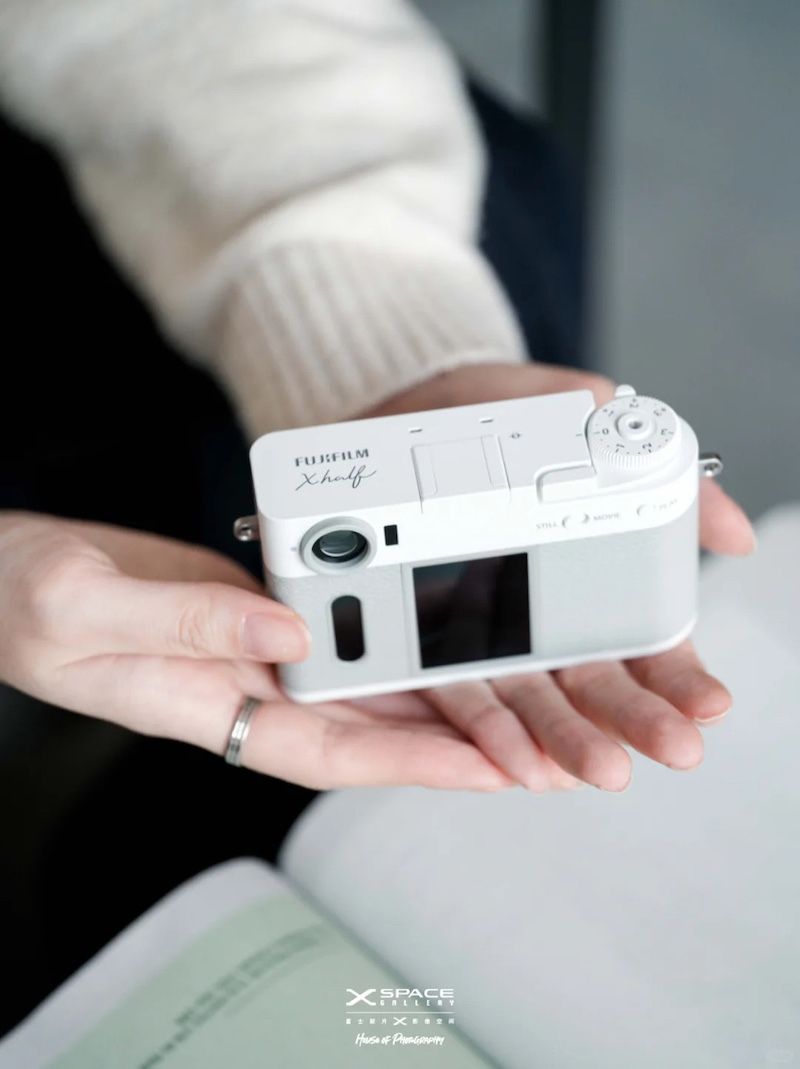 A person&rsquo;s hand holding a white Fujifilm X Half camera, with a soft-focus background of a book and indoor setting