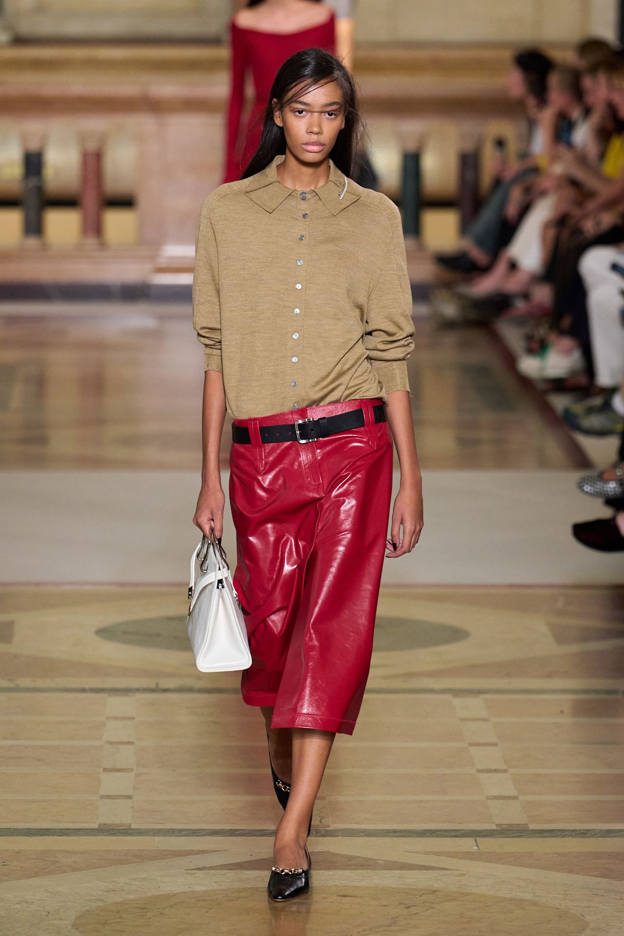 Layering staples for spring 2026 is shown in a photo from Tory Burch's runway show, with a model walking while wearing a tan polo layered under a tan cardigan styled with a red leather low-slung pencil skirt paired with a black belt, black pointed heels, and a white handbag