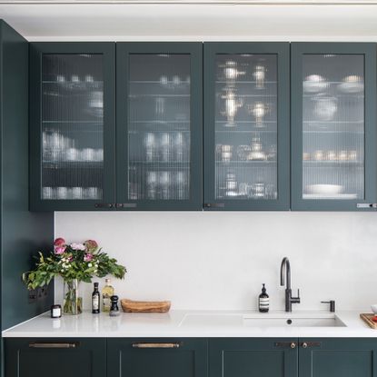 kitchen with white worktop and sink with a vase of flowers and dark green glass fronted cabinets