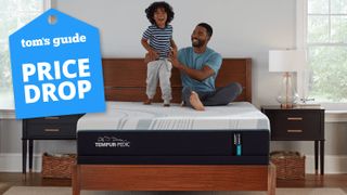 The Tempur-Pedic Tempur-Adapt mattress on a bed frame in a bedroom, a man and his young sun sitting on top of it, smiling. A Tom&#039;s Guide price drop deals graphic (right)