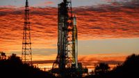 An orange rocket with white side booster stands next to the launch tower during a colorful sunrise.