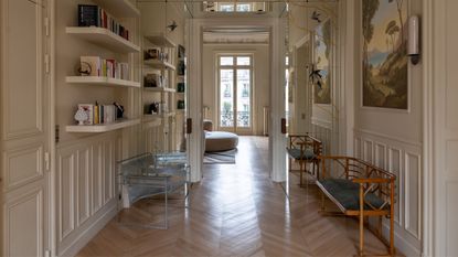 A grand hallway with off-white walls, paneling, and open shelving with seating benches along either side of the walls and paintings along the right-hand side