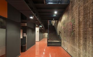 View from inside a corridor at the Cultural Centre, Dordrecht, rustic brick wall to the right, grey and orange partition wall to the left, doorways, black stairwell, orange gloss floor, ceiling spotlights